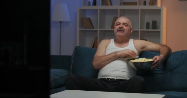 Wieczorem mężczyzna ogląda telewizję. Siedzi na kanapie. Jedzenie popcornu i kręcenie głową.. — Wideo stockowe