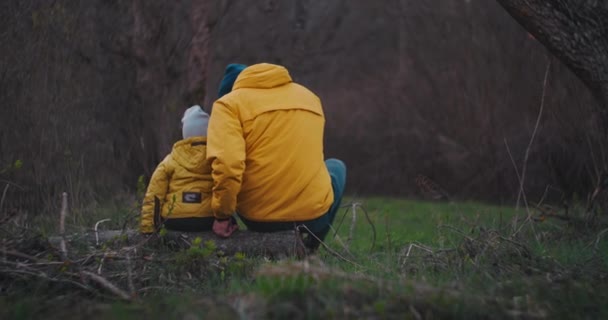 お父さんは芝生の上で息子の隣に座って話をして肩で抱きかかえる。黄色のジャケットで秋の森の中を歩く。カバーショット — ストック動画