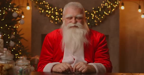 Papai Noel está sentado a uma mesa em uma bela sala de estar decorada para um feliz Natal. Papai Noel sorriu e acenou de acordo. — Vídeo de Stock