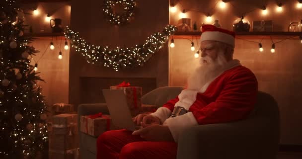 Santa Claus sits on a soft sofa in a beautifully decorated living room with lights and garlands. He holds a laptop on his lap and types text with one hand. — Stock Video