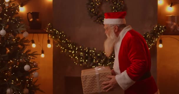 Santa Claus jde k vánočnímu stromečku a dává velkou krásnou krabici s dárkem pro poslušné dítě. — Stock video