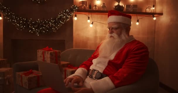 Święty Mikołaj siedzi na wygodnej sofie, trzymając laptopa na kolanach i pisząc na nim. Piękny salon urządzony na spotkanie Wesołych Świąt i Szczęśliwego Nowego Roku — Wideo stockowe