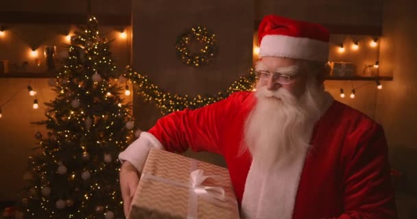 Le Père Noël se tient dans le salon à côté d'un sapin de Noël, puis prend une boîte avec un cadeau pour un enfant obéissant sous son aisselle pour un Joyeux Noël. Regarde la caméra et sourit. — Video