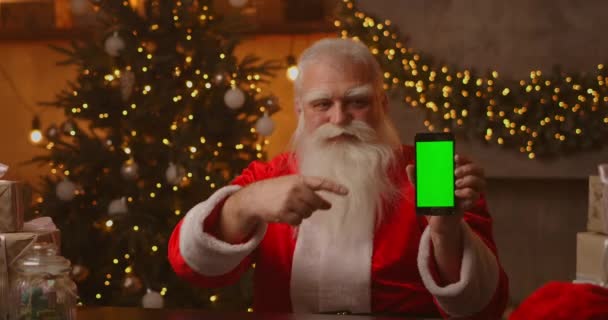 Kind Santa Claus sostiene un teléfono inteligente con una pantalla verde. Indica la pantalla del smartphone. Se sienta en una hermosa habitación decorada para una feliz Navidad. — Vídeos de Stock