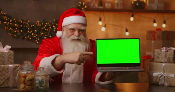 Tomten håller i en laptop med grön skärm. Indikerar skärmen på laptopen. Sitter i ett vackert rum inrett för en god jul. — Stockvideo