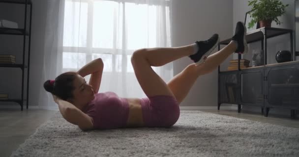 Vrouw is thuis aan het trainen, doet crunches in de woonkamer, thuis fitness in appartement, sportvrouw doet fysieke oefeningen voor buikspieren — Stockvideo