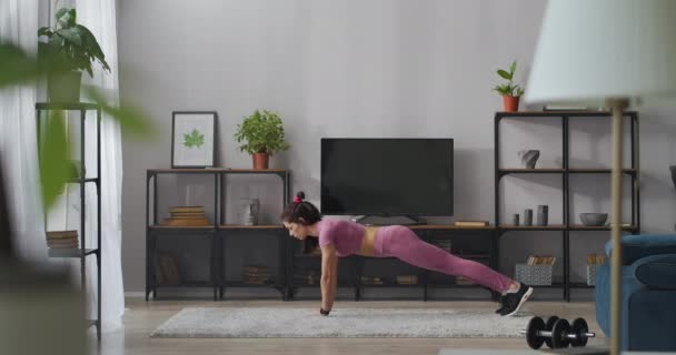 Спортсменка стоїть в дошці в кімнаті і піднімає ноги вгору, тренується вдома, зберігаючи пристосованість і хорошу форму тіла — стокове відео