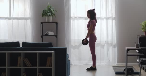 Esportista morena está treinando com halteres no quarto, de pé contra grande janela, inclinando-se para a frente — Vídeo de Stock