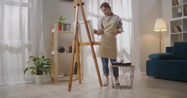 Giovane donna vestita grembiule sta lavorando in laboratorio artistico in appartamento, disegno e pittura con tinte e pennello su tela — Video Stock