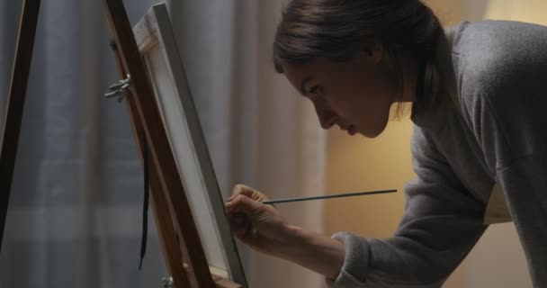 Жінка створює твори мистецтва на полотні, використовує барвники та пензлик, малює ввечері вдома, дивлячись на картину — стокове відео