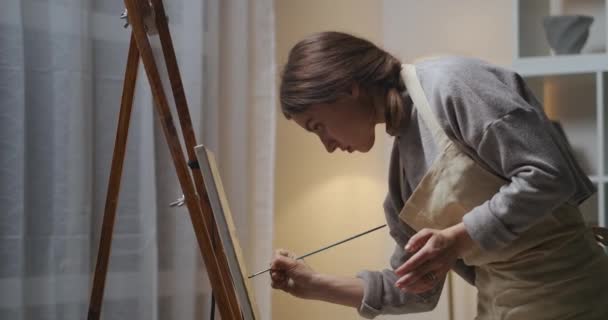 Hezká žena nosí zástěru a kreslí obraz olejovými barvami, večer pracuje v domácím uměleckém studiu — Stock video