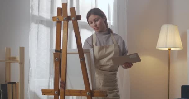 Giovane bella donna sta disegnando immagine in soggiorno, in piedi vicino cavalletto con tela, artista femminile sta lavorando — Video Stock