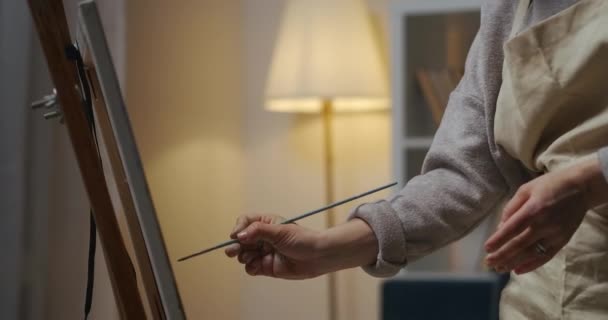 Жінка-художник малює вдома ввечері, крупним планом дивиться на руки пензлем, наносить барвники на полотно — стокове відео