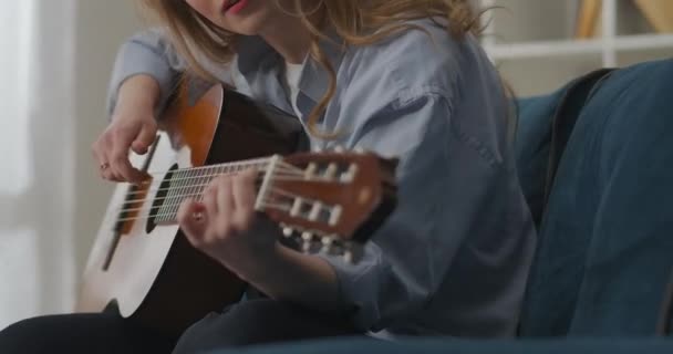 Giovane musicista donna sta pizzicando corde di chitarra, seduta sul divano in casa, amante della musica country donna, dettagli girato — Video Stock