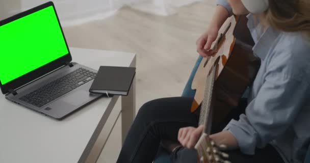 Mujer está aprendiendo a tocar la guitarra por tutorial en línea, escuchando por auriculares, tecnología de pantalla verde en el portátil — Vídeo de stock
