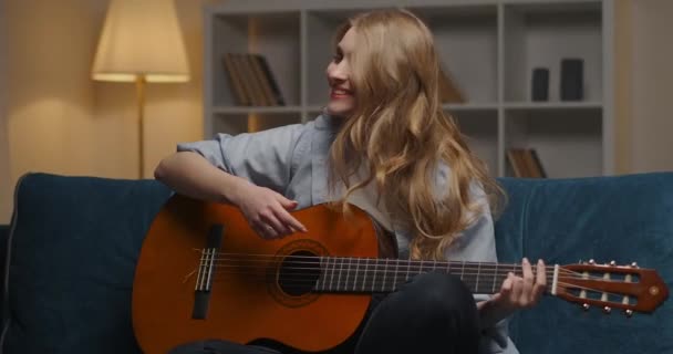 Alegre joven con la guitarra se divierte en casa por la noche, sentado solo en acogedor apartamento y disfrutando de la música — Vídeo de stock