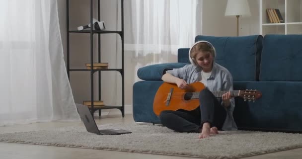 Prove musicali online, la donna sta suonando la chitarra e ascoltando musica con le cuffie, guardando sul display del computer portatile — Video Stock