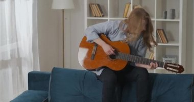 Acemi country şarkıcısı kadın evdeki koltuğun arkasında gitar çalıyor, müzikten zevk alıyor.