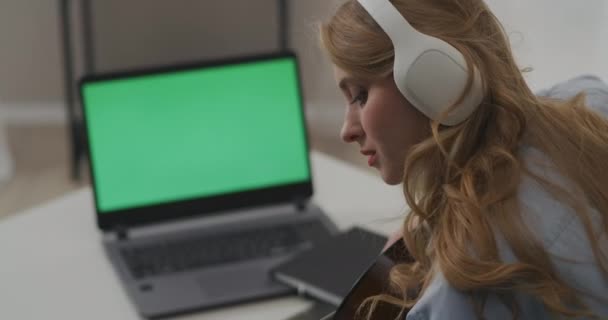 Country músico feminino está tocando guitarra em casa, sentado na frente do laptop aberto com tela verde, streaming on-line no chat de vídeo — Vídeo de Stock