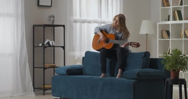 Romantische jonge vrouw zit met gitaar op de rug van de bank in gezellige woonkamer en het spelen van muziek, liefdeslied — Stockvideo