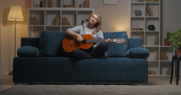 행복 한 아름다운 여성은 음악을 즐기고, 아파트 소파에 앉아 저녁에 집에서 기타를 치고 있습니다. — 비디오