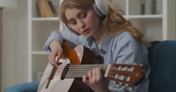 Jonge mooie vrouw met koptelefoon op het hoofd is studeren om gitaar te spelen, alleen in appartement in het weekend — Stockvideo