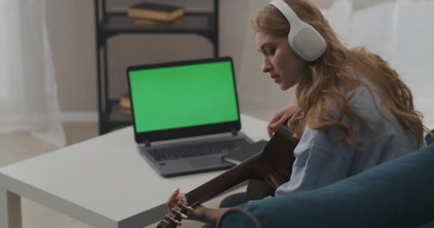 Hübsche Musikerin spielt Gitarre in der Wohnung, grüner Bildschirm des Laptops auf dem Tisch im Zimmer, Online-Streaming — Stockvideo