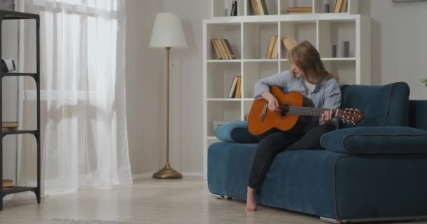 Joven bonita mujer está tocando la guitarra y cantando canción, pasar tiempo en fin de semana solo en su casa — Vídeos de Stock