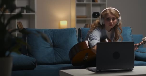 Teenie-Mädchen spielt Gitarre, liest Notizen und Tutorial im Internet auf Notebook am Abend, verbringt Freizeit und lernt online — Stockvideo