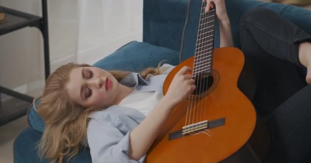 美しい女性はアパートのソファに横になってアコースティックギターを弾いたりロマンチックな気分で音楽を楽しんでいます — ストック動画