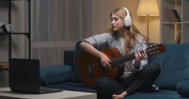 Mujer joven despreocupada está pasando la noche en casa, aprendiendo a tocar la guitarra, sentado en el sofá con auriculares y ordenador portátil — Vídeo de stock