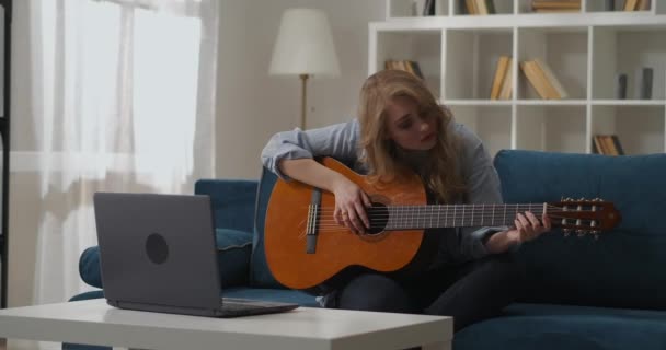 Jeune femme blonde joue de la guitare assis à la maison sur le canapé, apprendre à jouer seul, la musique et le passe-temps — Video
