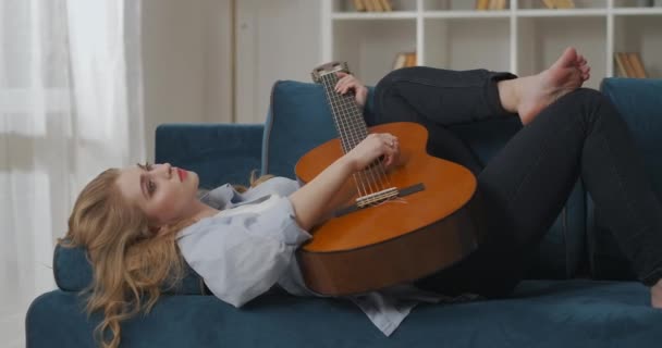 Începător country muzician femeie este culcat pe canapea la domiciliu și cântând muzică pe chitară, talentată și doamnă inspirată — Videoclip de stoc