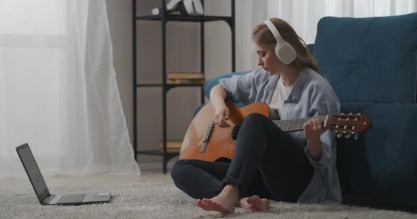 Mujer está jugando música sentada en el suelo en la habitación, utilizando video guía de autoayuda para tocar la guitarra en el ordenador portátil — Vídeos de Stock