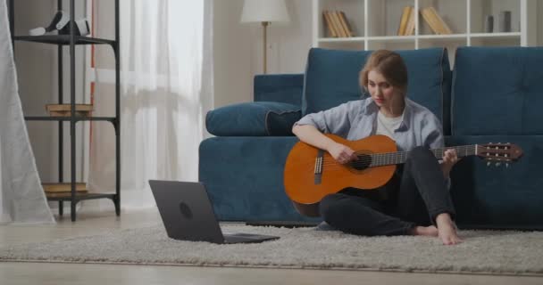 Mooie dame zit op de vloer in appartement en spelen lied op gitaar, leren via internet, verre muziek klasse — Stockvideo