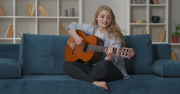 Donna spensierata sta suonando la chitarra e cantando, seduto sul divano da solo a casa, divertimento e felicità nel fine settimana — Video Stock