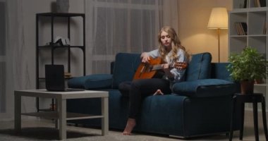 Genç kadın gitarda hüzünlü bir aşk şarkısı çalıyor, akşam oturma odasında oturuyor, romantik bir ruh hali, müzik ve hobi.