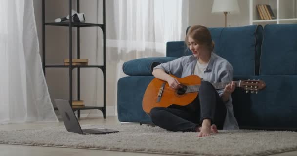 Fin de semana tranquilo en casa con la guitarra, la mujer está aprendiendo a jugar, ver tutorial en el ordenador portátil, educación en línea, pasatiempo — Vídeos de Stock