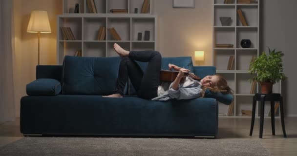 Relax at home at evening, woman is lying with guitar on couch in cozy apartment and playing calm song — Stock Video