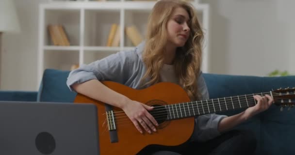 Young female beginner musician is learning accords on guitar, playing on strings, sitting on couch in apartment — Stock Video