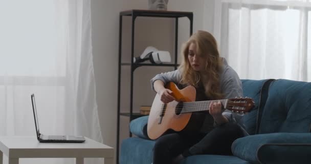 Giovane musicista femminile sta suonando la chitarra, seduto a casa e la visualizzazione di video tutorial sul computer portatile, imparare online, hobby — Video Stock