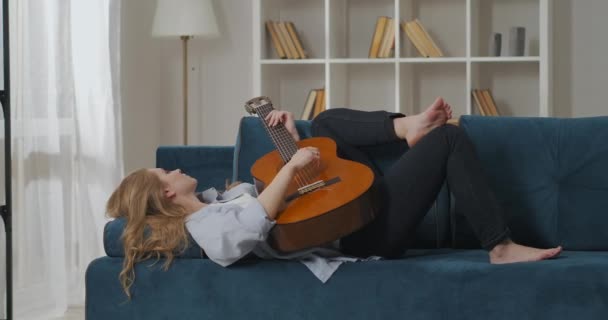 Беззаботная романтическая женщина лежит на диване дома и играет на гитаре, наслаждаясь музыкой и отдыхом в квартире по выходным — стоковое видео