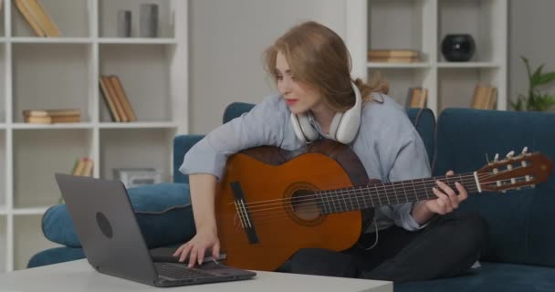 여자는 인터넷에서 기타를 연주하는 방법에 대한 정보를 찾고 있습니다. 노트북에서 기타를 들고 줄을 뽑고 스스로 교육하는 것이죠. — 비디오