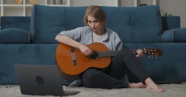 Donna gioiosa sta giocando la chitarra seduta a casa sul pavimento, imparare una nuova canzone con le istruzioni in internet, guardando sul display del notebook — Video Stock