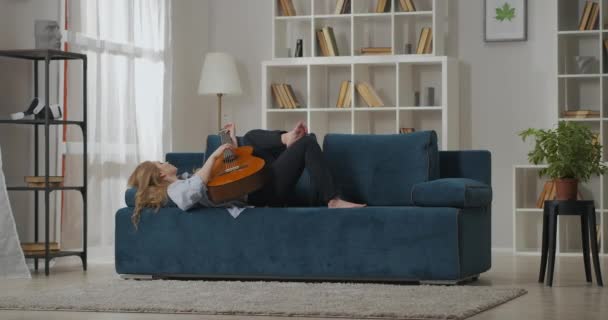 Νεαρή γυναίκα ξεκουράζεται στο σπίτι το Σαββατοκύριακο, ξαπλωμένη στον καναπέ στο σαλόνι και παίζοντας κιθάρα, απολαμβάνοντας μουσική και αγαπημένο τραγούδι — Αρχείο Βίντεο