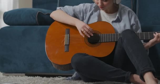 Cântă la chitară singură acasă, tânăra face exerciții la chitară, ascultă melodia prin căști — Videoclip de stoc