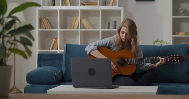 young sad woman is playing guitar at home, viewing notes on screen of laptop, learning to play, composing music