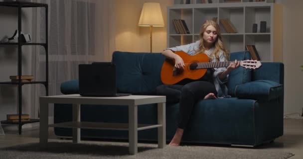 젊고 아름다운 여성이 저녁에 집에서 기타를 치고, 휴식과 여가를 즐기고, 인터넷으로 연주하는 법을 배우고 있습니다. — 비디오