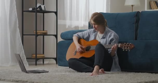 Jonge vrouwelijke muzikant is het leren van nieuwe akkoorden op gitaar, zitten op de vloer in appartement en het bekijken van tutorials op internet door laptop — Stockvideo