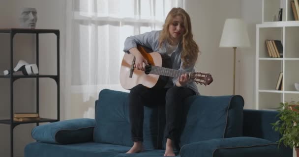 Felice donna sta cantando canzone e suonare la chitarra a casa la domenica, seduto sul retro del divano e divertirsi — Video Stock